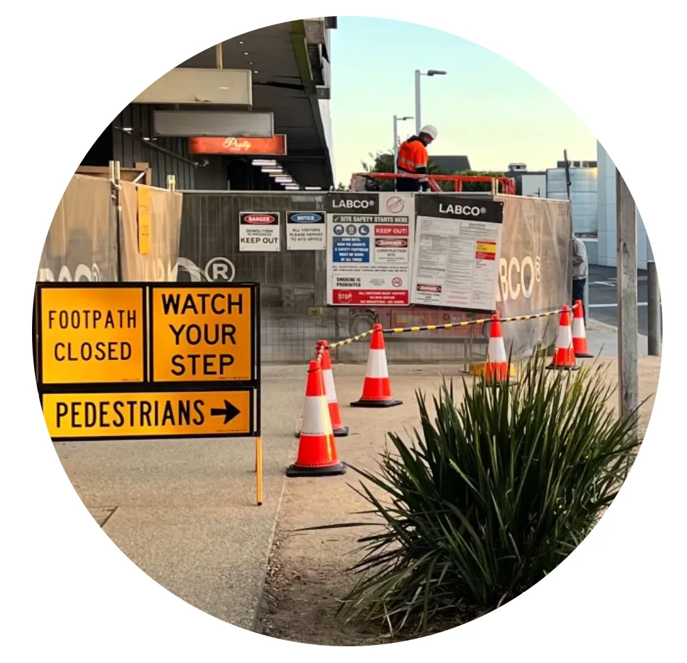 Construction site with “FOOTPATH CLOSED,” “WATCH YOUR STEP,” and “PEDESTRIANS →” signs, traffic cones, and a hi-vis worker behind safety fencing.